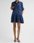 A woman modeling a short-sleeve blue tiered shirt dress with a textured crinkle fabric, paired with black strappy heels against a light gray studio background.