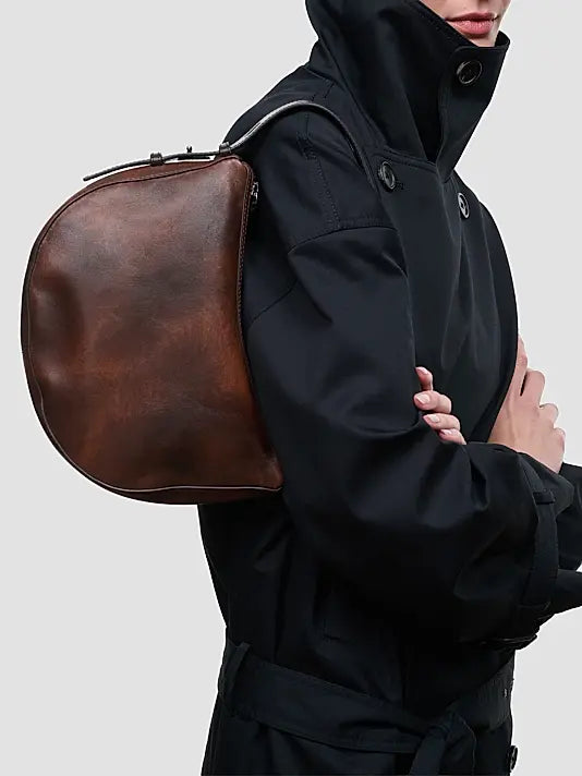 A person wearing a black hooded jacket carries a large, semi-circular brown leather shoulder bag against their back, shown against a plain light gray background.