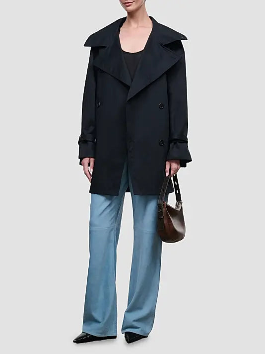 A model wearing a black double-breasted trench coat over a black top, paired with light blue wide-leg trousers and holding a brown leather handbag, set against a solid light gray background.
