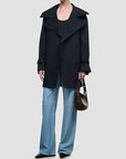 A model wearing a black double-breasted trench coat over a black top, paired with light blue wide-leg trousers and holding a brown leather handbag, set against a solid light gray background.