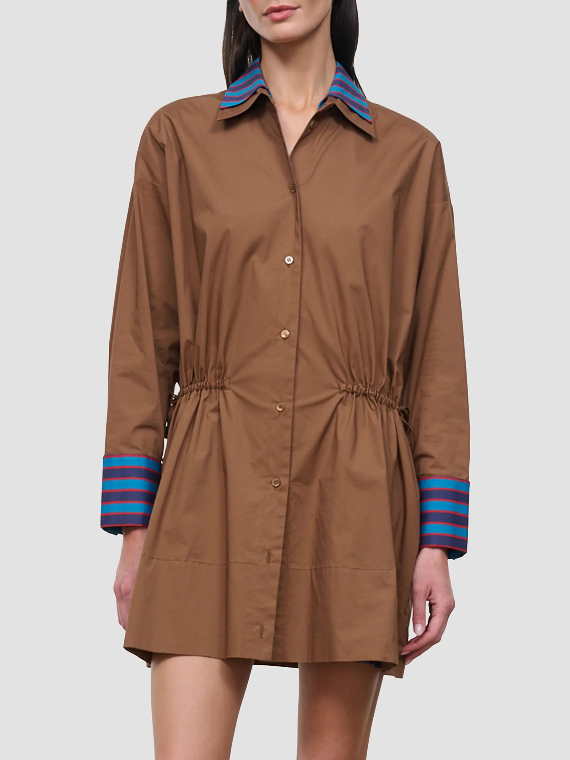 A model wears a brown long-sleeve shirt dress with a pointed collar, button front, gathered waist, and striped cuffs in blue, purple, and red, set against a solid light gray background.