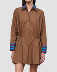 A model wears a brown long-sleeve shirt dress with a pointed collar, button front, gathered waist, and striped cuffs in blue, purple, and red, set against a solid light gray background.