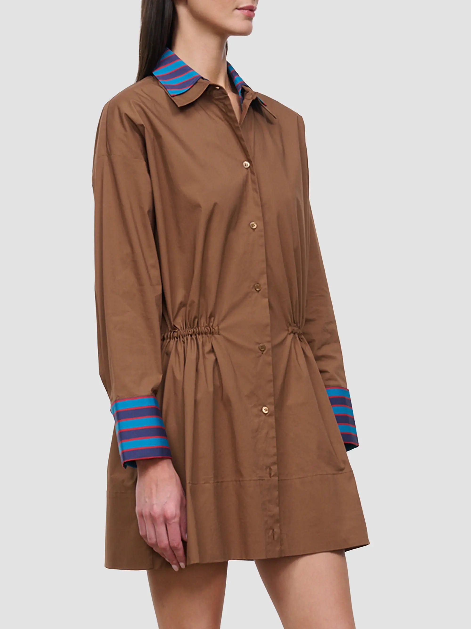 A woman wearing a brown long-sleeve shirt dress with a blue and red striped collar and cuffs, shown against a light gray studio background.