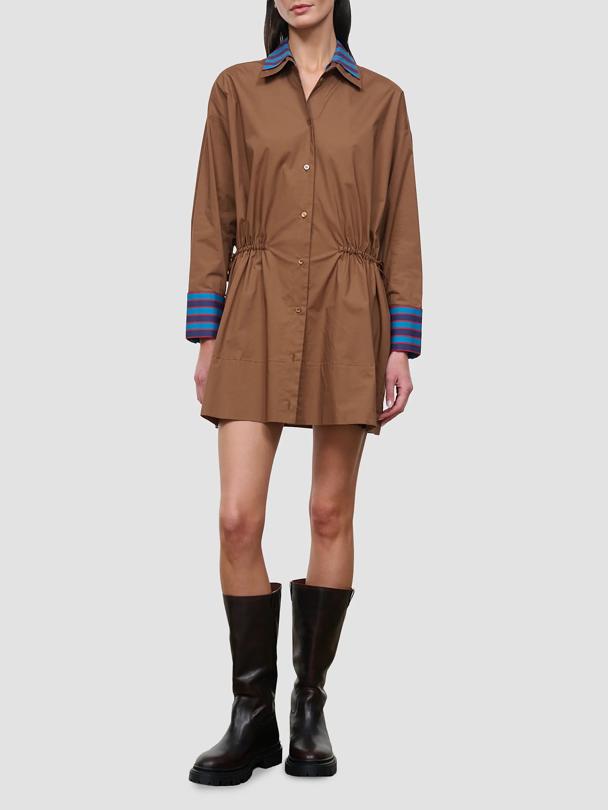 A model wears a brown long-sleeve shirt dress with a blue striped collar and cuffs, paired with black boots, against a solid gray background.