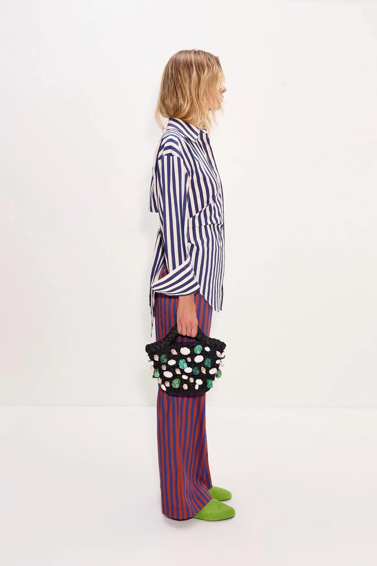 Person wearing a striped shirt and colorful pants holding a polka dot bag on a white background, side
