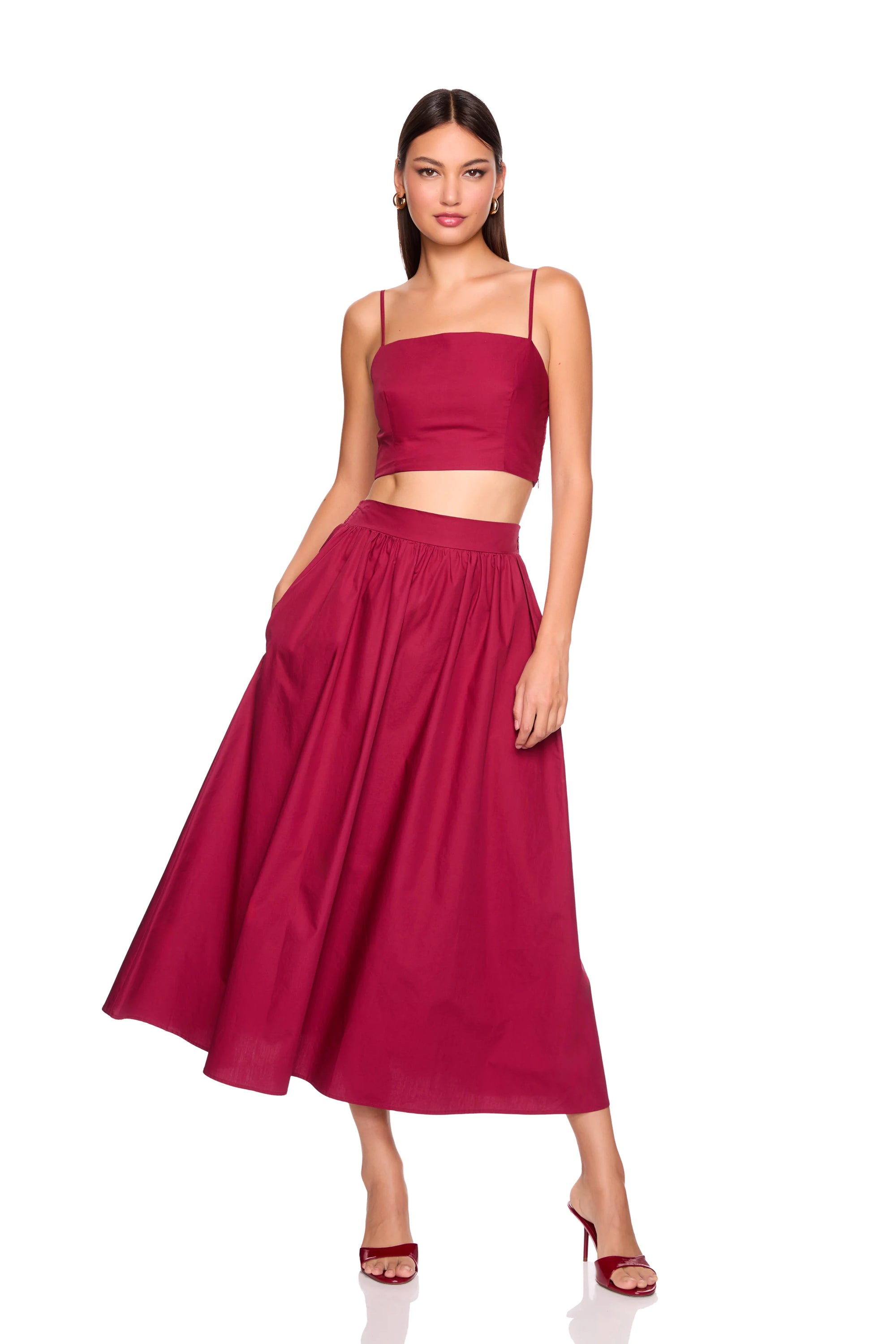 A woman modeling a two-piece outfit consisting of a deep magenta crop top with spaghetti straps and a matching high-waisted pleated midi skirt, paired with matching heeled sandals against a plain white background.