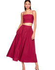A woman modeling a two-piece outfit consisting of a deep magenta crop top with spaghetti straps and a matching high-waisted pleated midi skirt, paired with matching heeled sandals against a plain white background.