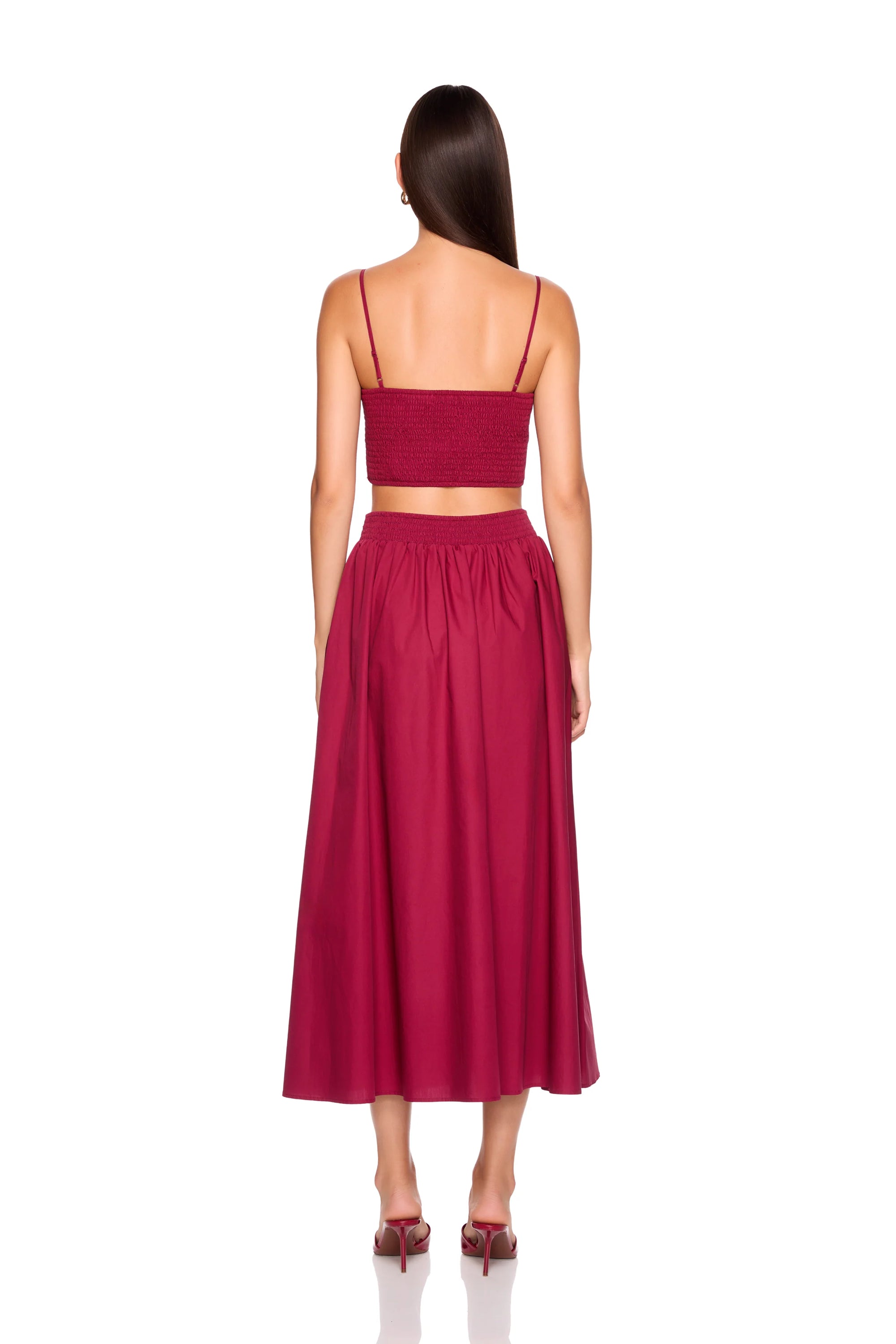 A woman viewed from the back wearing a matching two-piece outfit consisting of a deep magenta spaghetti-strap crop top and a high-waisted pleated midi skirt, paired with dark brown heels against a plain white background.