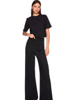 A woman modeling a black short-sleeve mock-neck top and matching high-waisted black wide-leg pants against a plain white background.