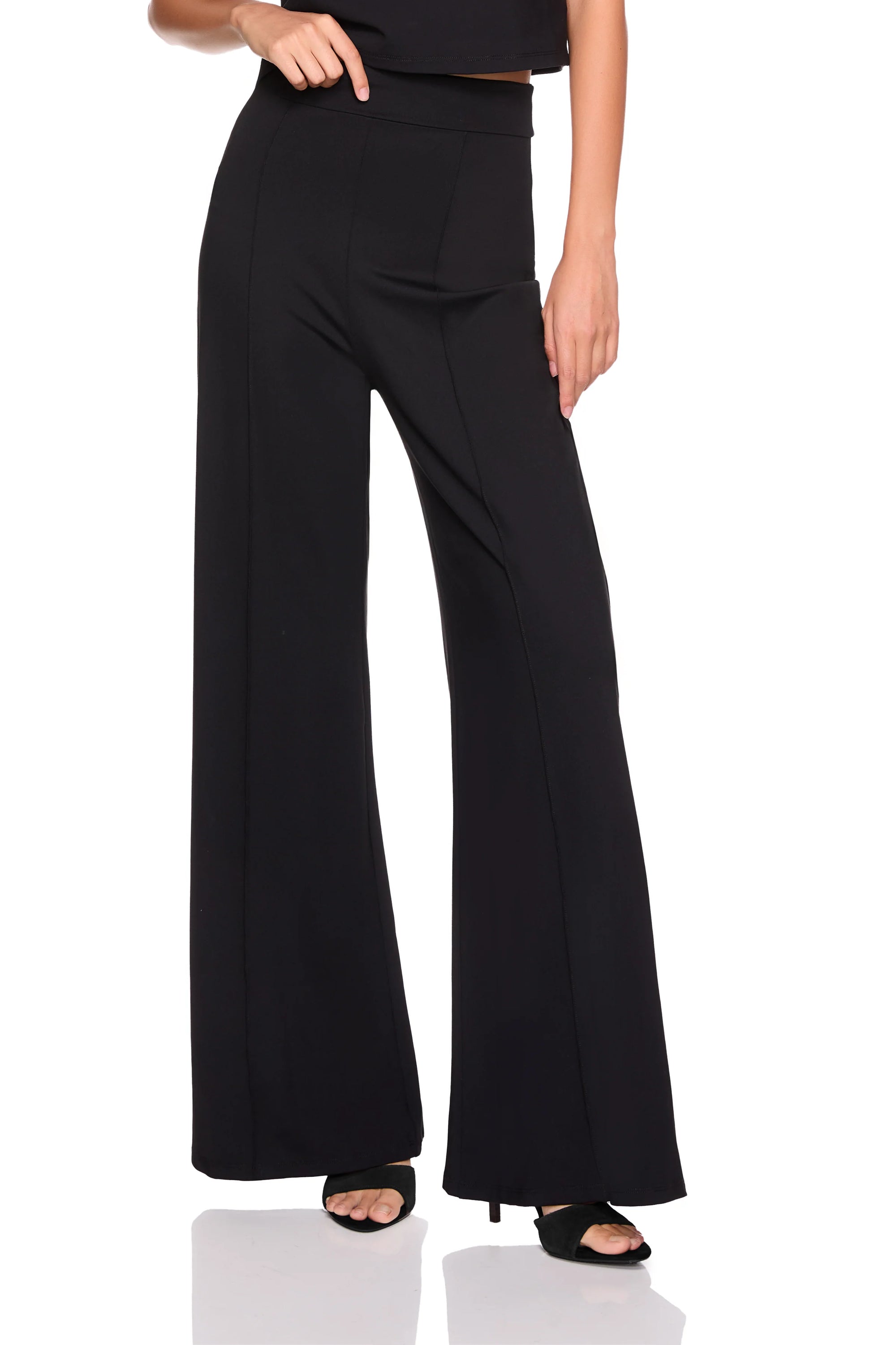 A model wearing black wide-leg high-waisted pants with a front welt pocket, paired with black heeled sandals, shown from the waist down on a white background.
