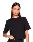 A woman modeling a black short-sleeve crop top with a relaxed, slightly oversized fit, paired with black bottoms, against a solid white background.