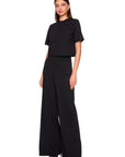 A woman modeling a black two-piece outfit consisting of a short-sleeve cropped top and matching high-waisted wide-leg trousers, photographed against a plain white studio background.
