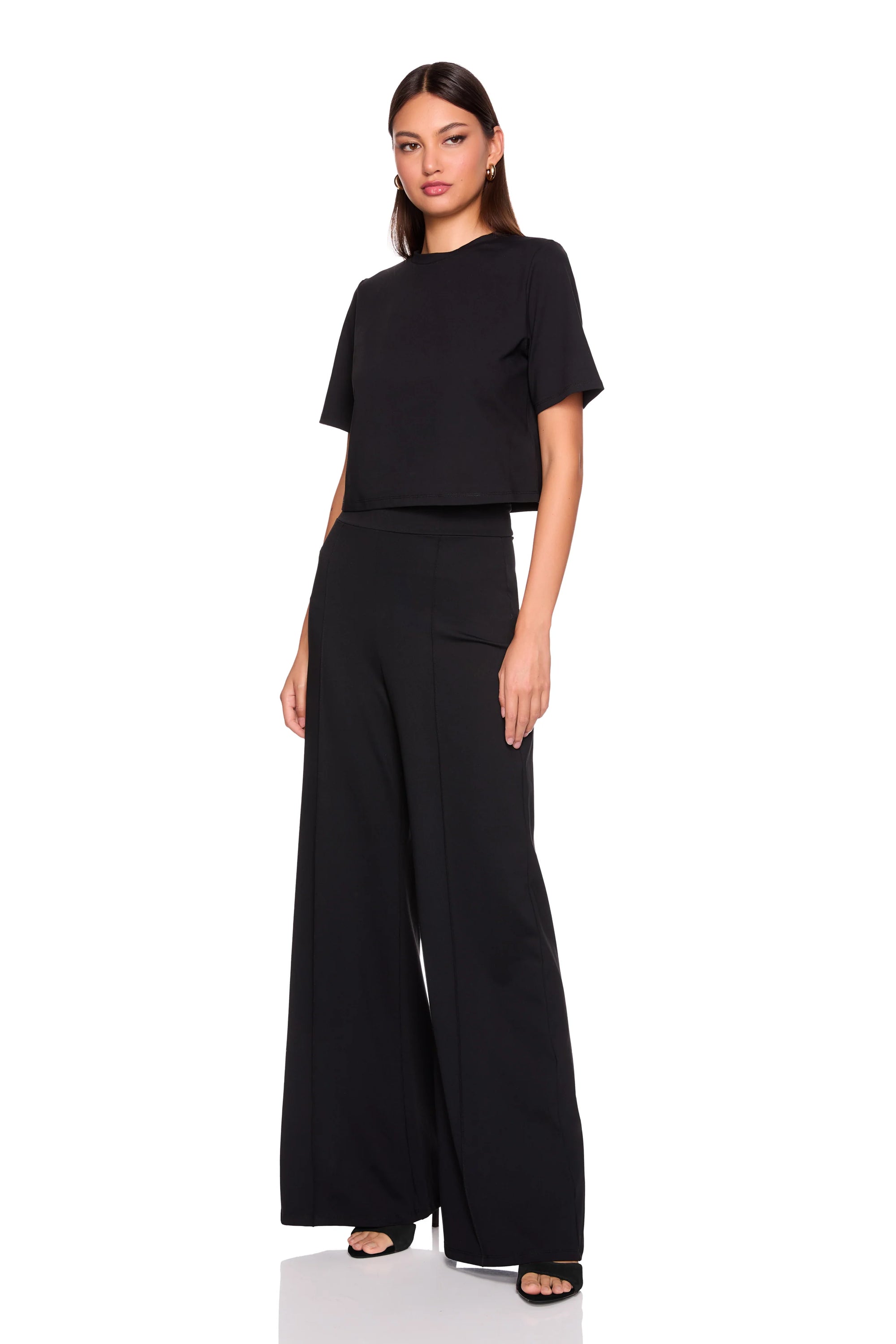 A woman modeling a black two-piece outfit consisting of a short-sleeve cropped top and matching high-waisted wide-leg trousers, photographed against a plain white studio background.