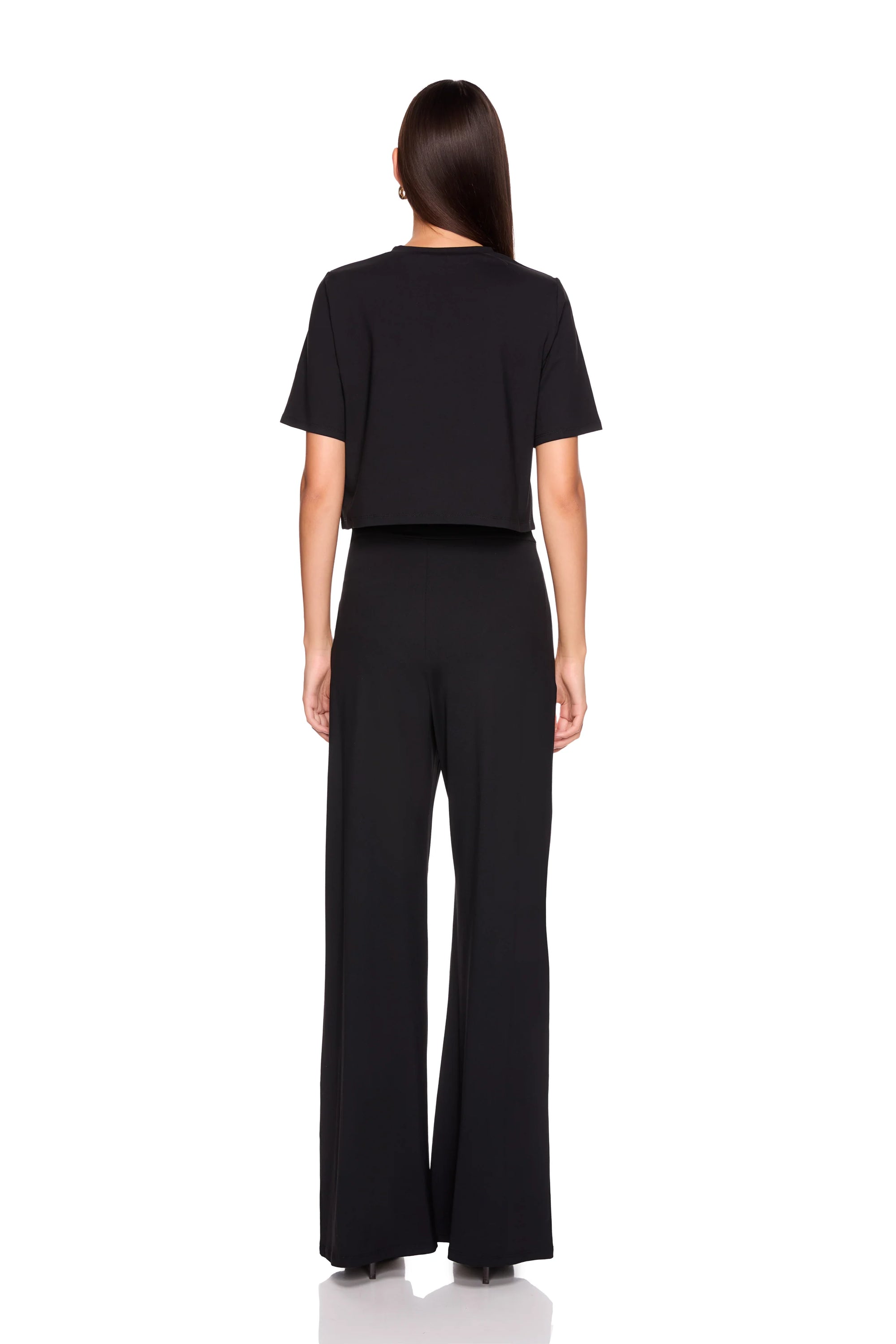 A woman viewed from the back wearing a black short-sleeve cropped top and matching black wide-leg pants against a plain white studio background.