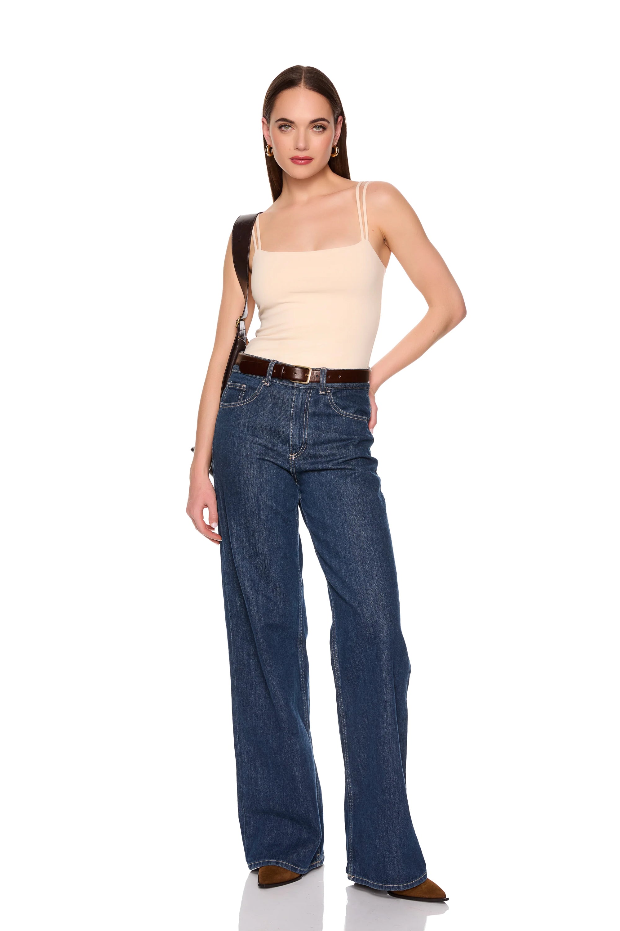 A woman modeling a pair of high-waisted, wide-leg blue jeans with a dark brown belt, paired with a beige camisole top and brown shoes, on a solid white background.