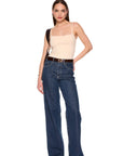 A woman modeling a pair of high-waisted, wide-leg blue jeans with a dark brown belt, paired with a beige camisole top and brown shoes, on a solid white background.