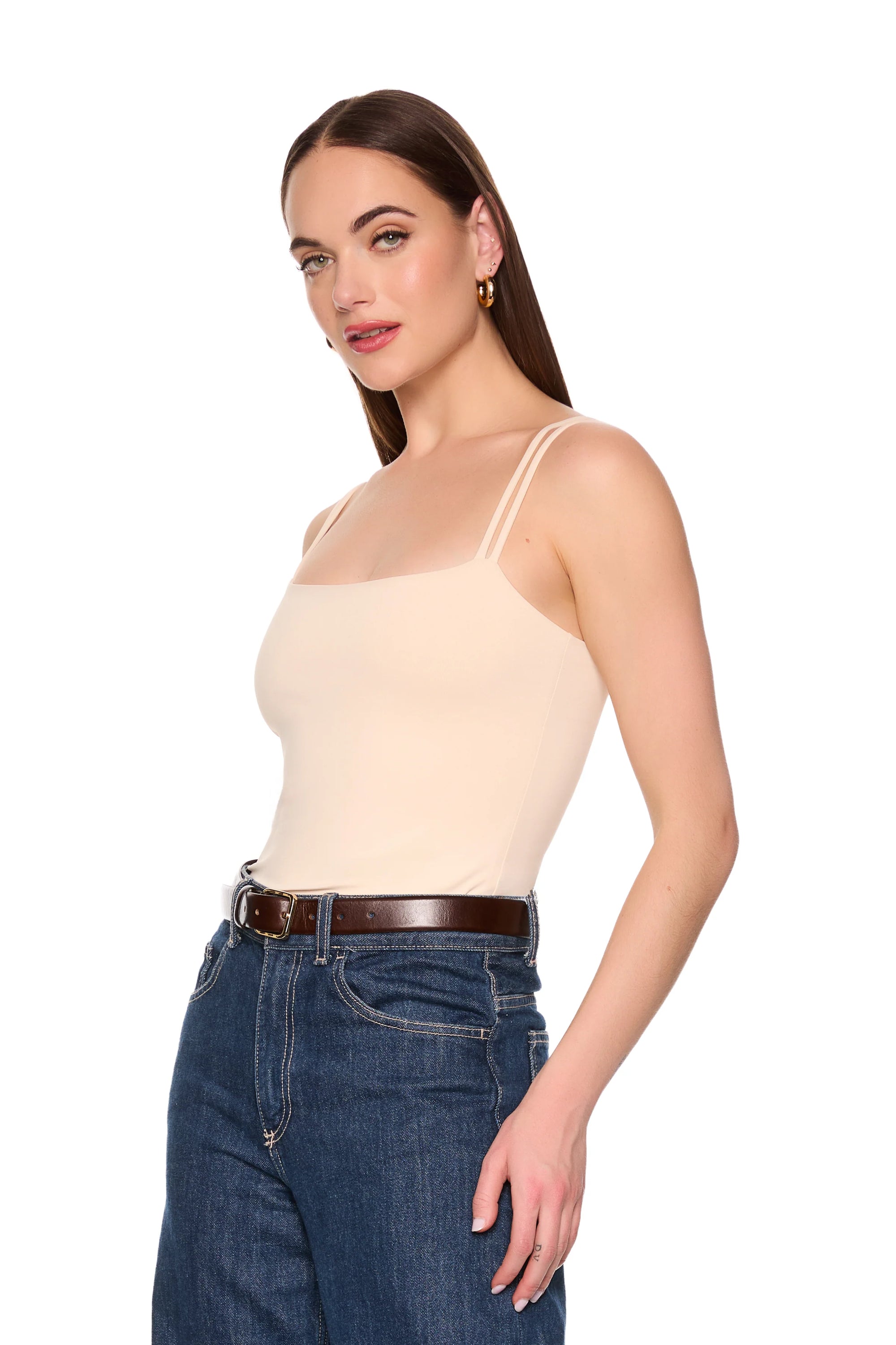 A woman modeling a beige spaghetti-strap camisole top paired with dark blue jeans and a brown belt, photographed against a solid white background.