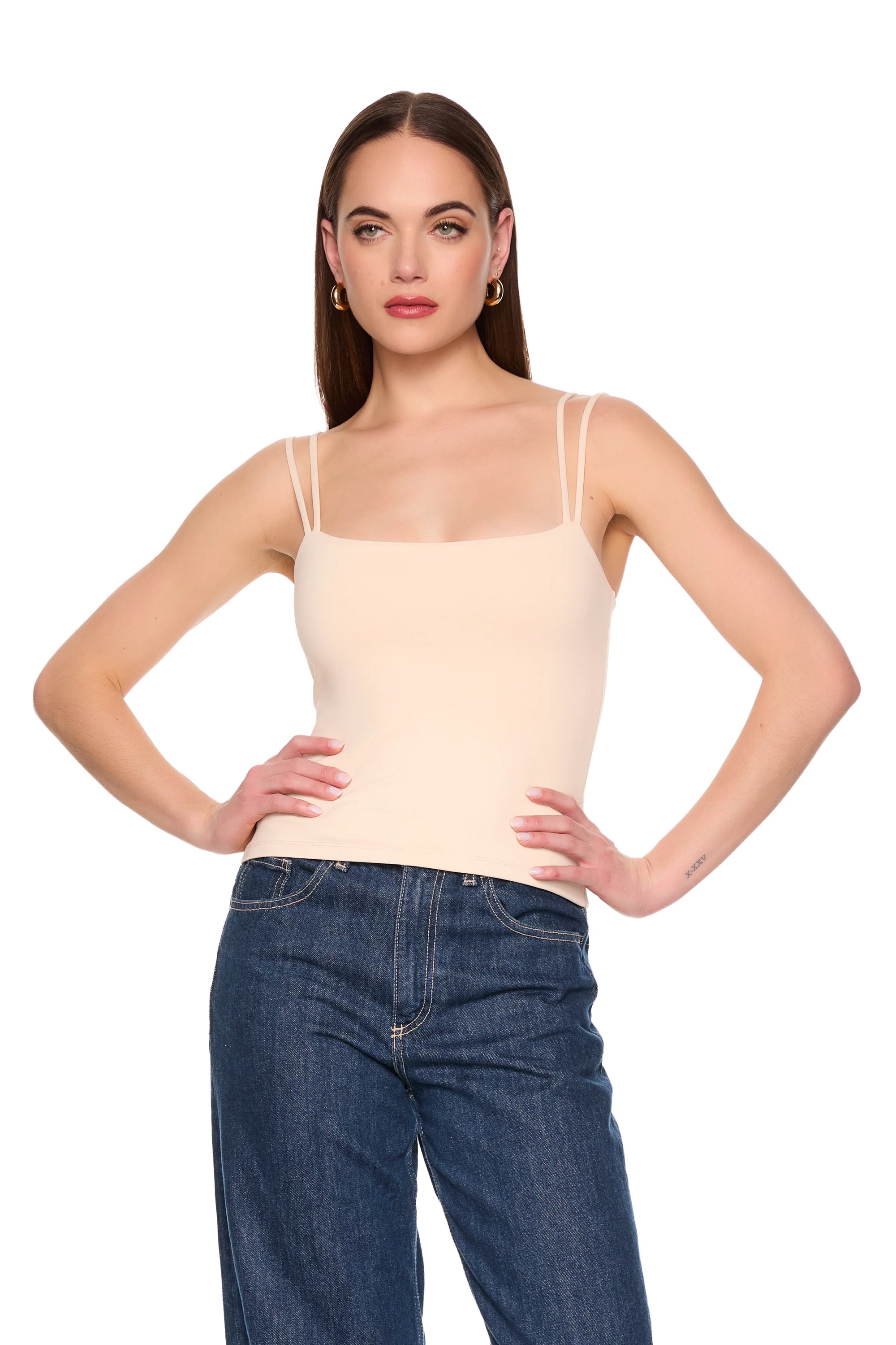A woman modeling a light beige spaghetti-strap camisole top paired with dark blue jeans, standing against a solid white background.