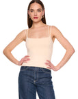 A woman modeling a light beige spaghetti-strap camisole top paired with dark blue jeans, standing against a solid white background.