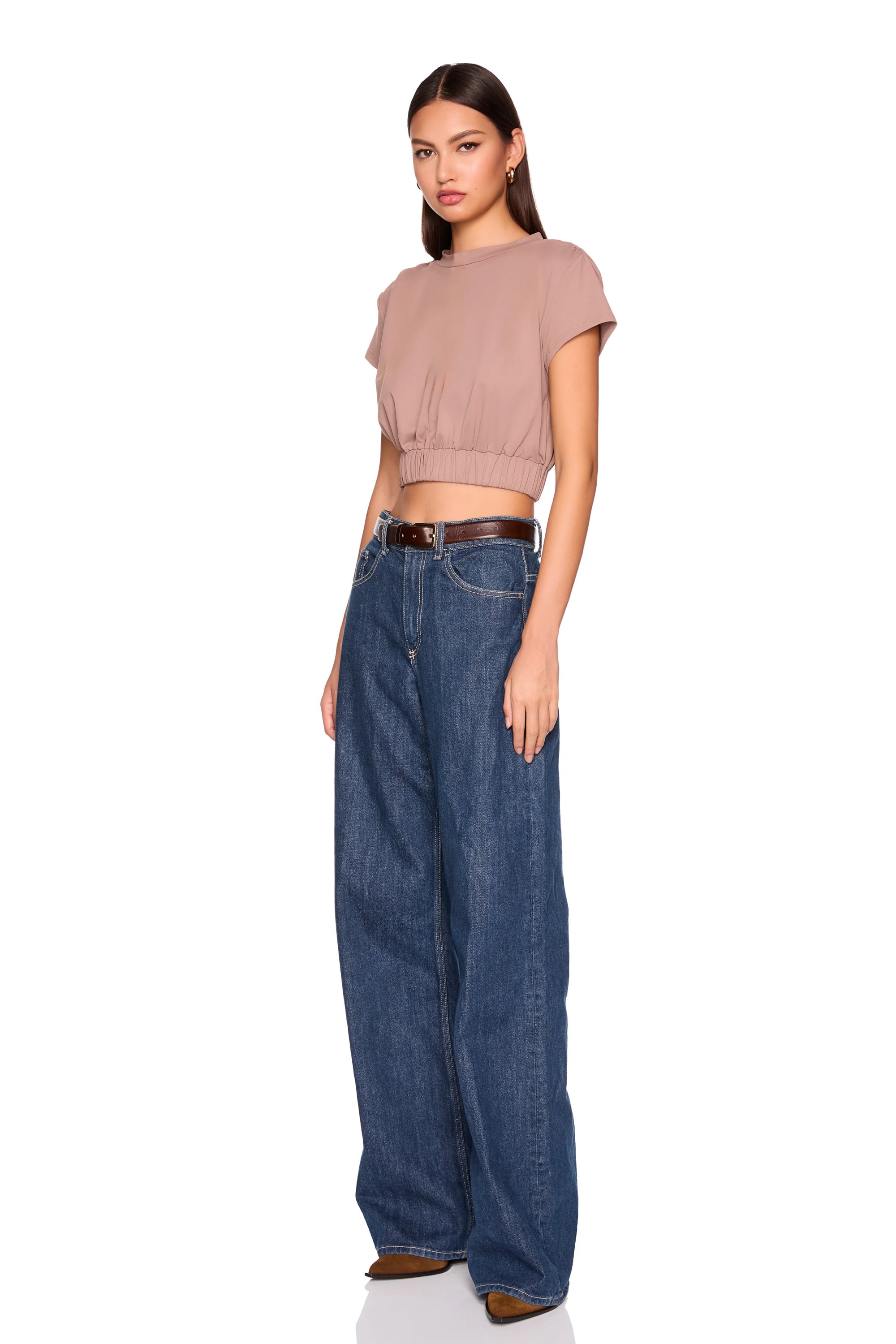 A model wearing a dusty rose short-sleeve crop top and high-waisted, wide-leg blue jeans with a brown belt, standing against a solid white background.