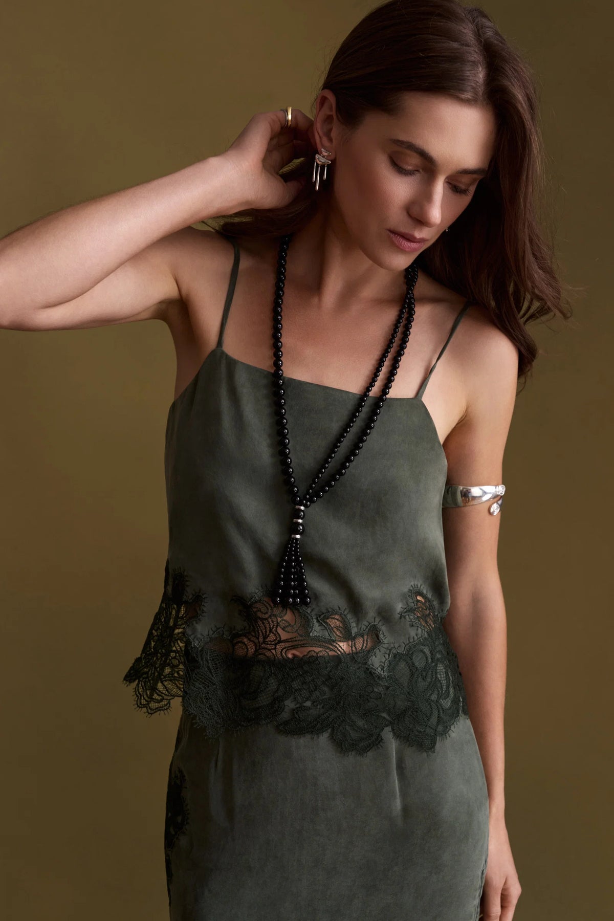 A woman wearing a dark olive green camisole dress with lace trim and a long black beaded tassel necklace, posed against a solid olive-brown background.
