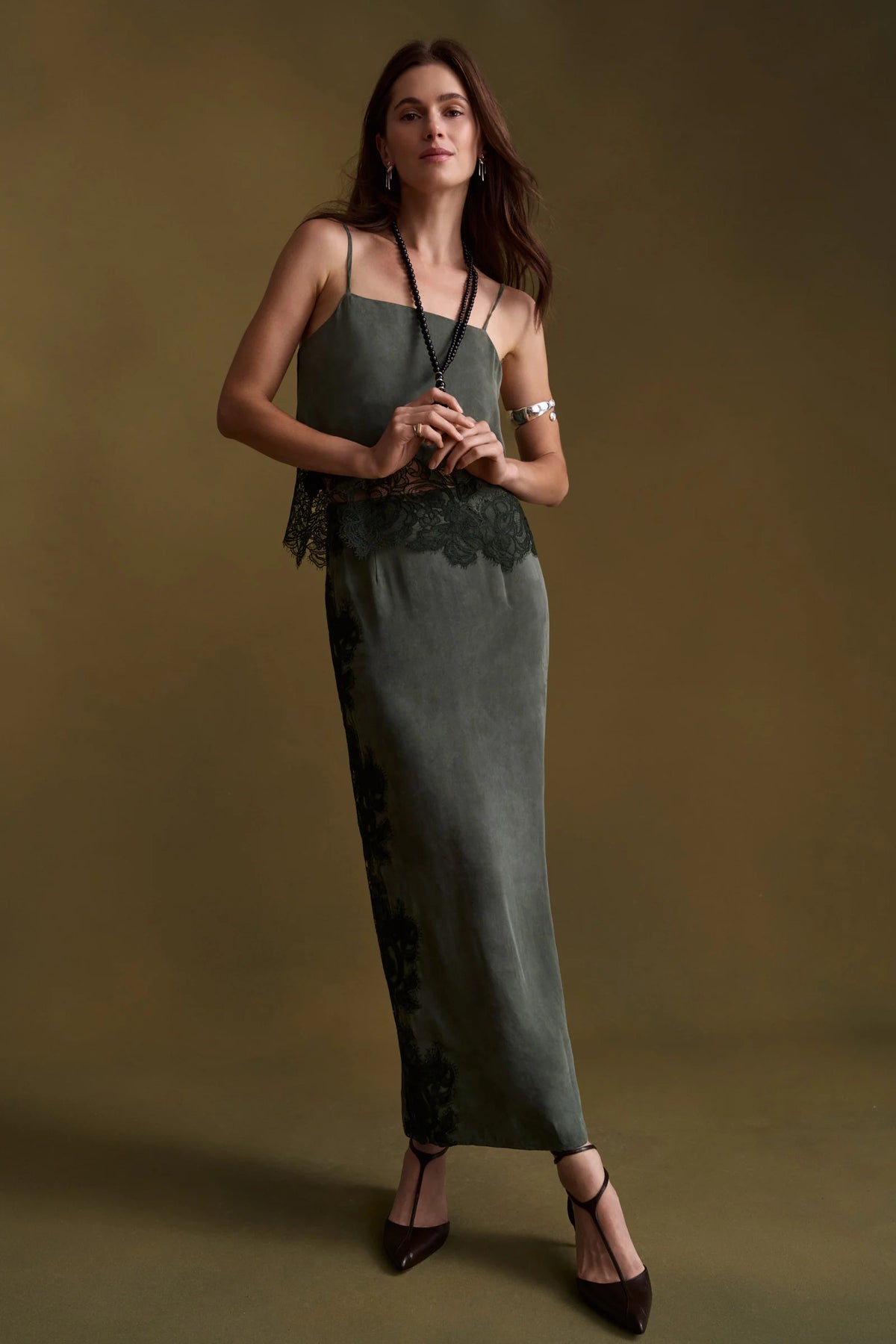 A woman modeling a dark olive green sleeveless set with lace trim at the bust and along the side, paired with black strappy heels, against a solid dark olive background.