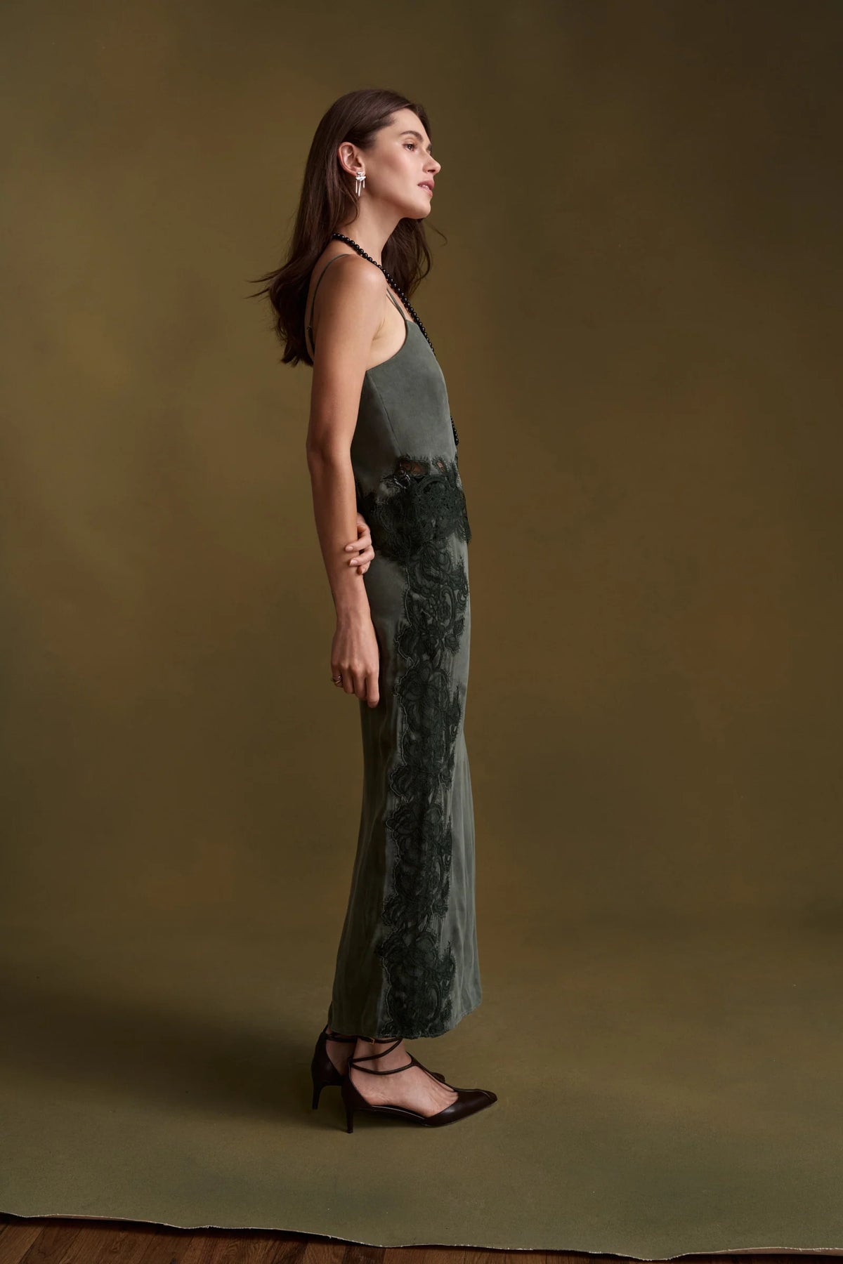 A woman stands in profile wearing a sleeveless, floor-length olive green set with a floral lace overlay on the skirt, posed against a solid olive-brown studio backdrop.