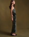 A woman stands in profile wearing a sleeveless, floor-length olive green set with floral lace overlay on the skirt, posed against a solid olive-brown studio backdrop.