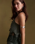 A woman wearing a dark olive green sleeveless dress with lace detailing and a wide silver cuff bracelet on her arm, posed against a solid olive-brown background.