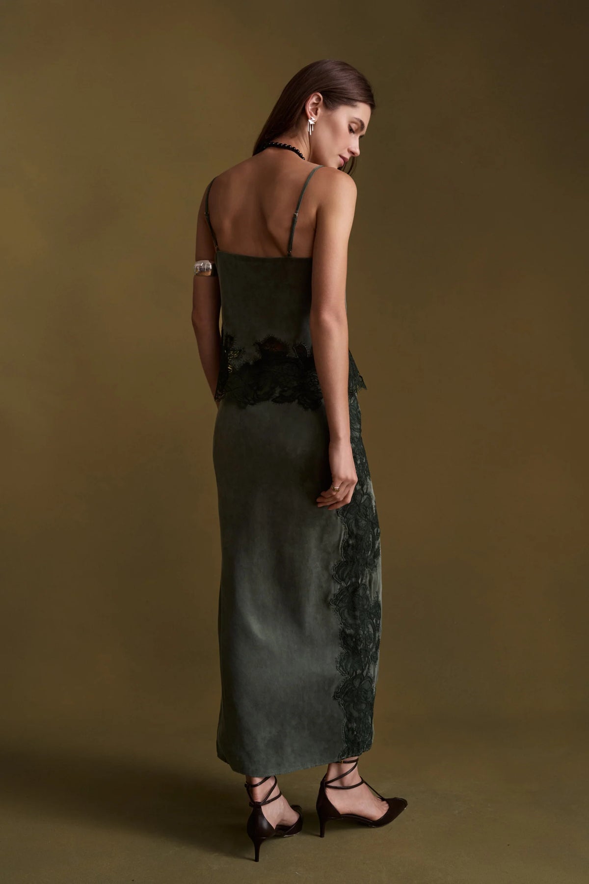 A woman wearing a dark green set with black lace trim, photographed from the back against a solid olive-brown studio background.