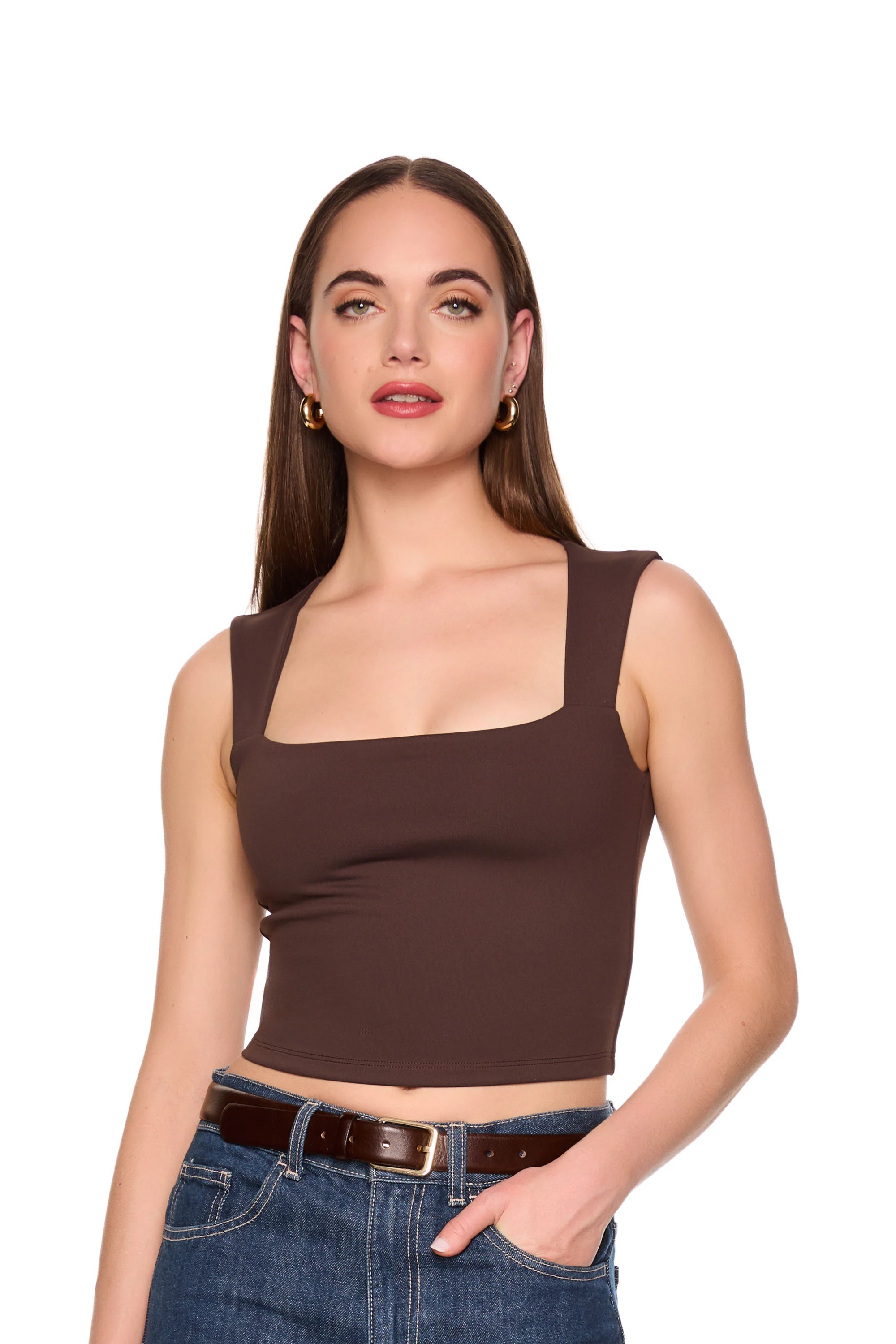 A woman modeling a dark brown square-neck crop top with wide straps, paired with blue jeans and a brown belt, against a solid white background.