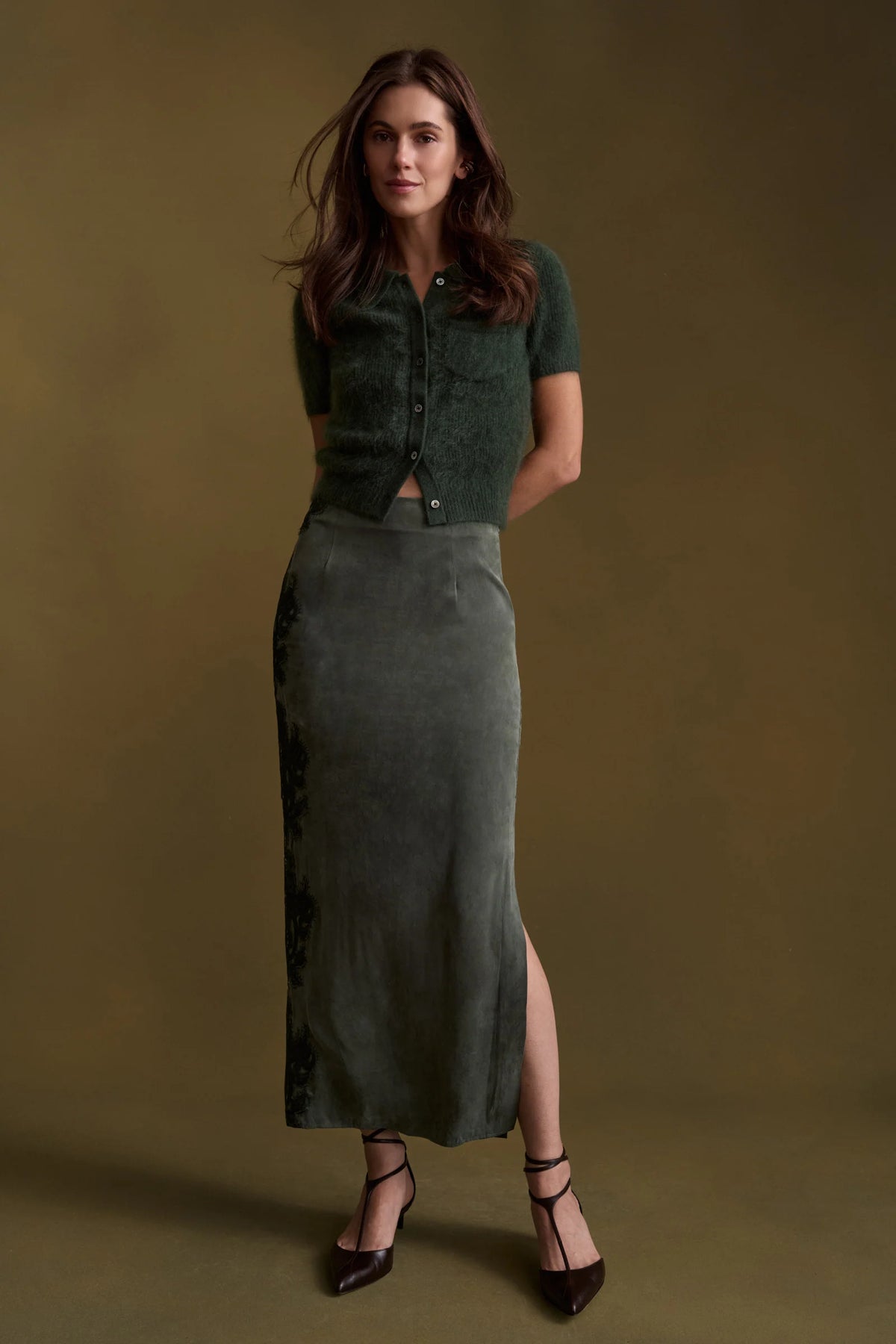 A woman stands against a warm olive-brown studio backdrop wearing a dark green short-sleeve cropped knit top with front buttons and a matching dark green midi skirt with a side slit, paired with black strappy heels.