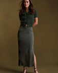 A woman stands against a warm olive-brown studio backdrop wearing a dark green short-sleeve cropped knit top with front buttons and a matching dark green midi skirt with a side slit, paired with black strappy heels.