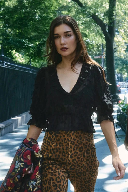 A woman walking outdoors on a tree-lined sidewalk wearing a black ruffled lace blouse and leopard-print pants, holding a floral garment in one hand.
