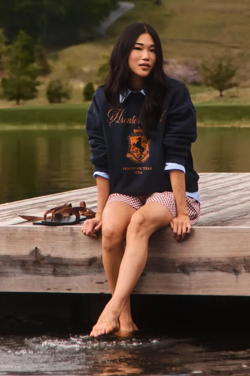 A woman sits barefoot on the edge of a wooden dock over water, wearing a navy blue sweatshirt over a light blue collared shirt, paired with red-and-white gingham shorts.