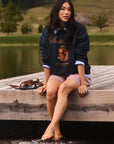 A woman sits barefoot on the edge of a wooden dock over water, wearing a navy blue sweatshirt over a light blue collared shirt, paired with red-and-white gingham shorts.