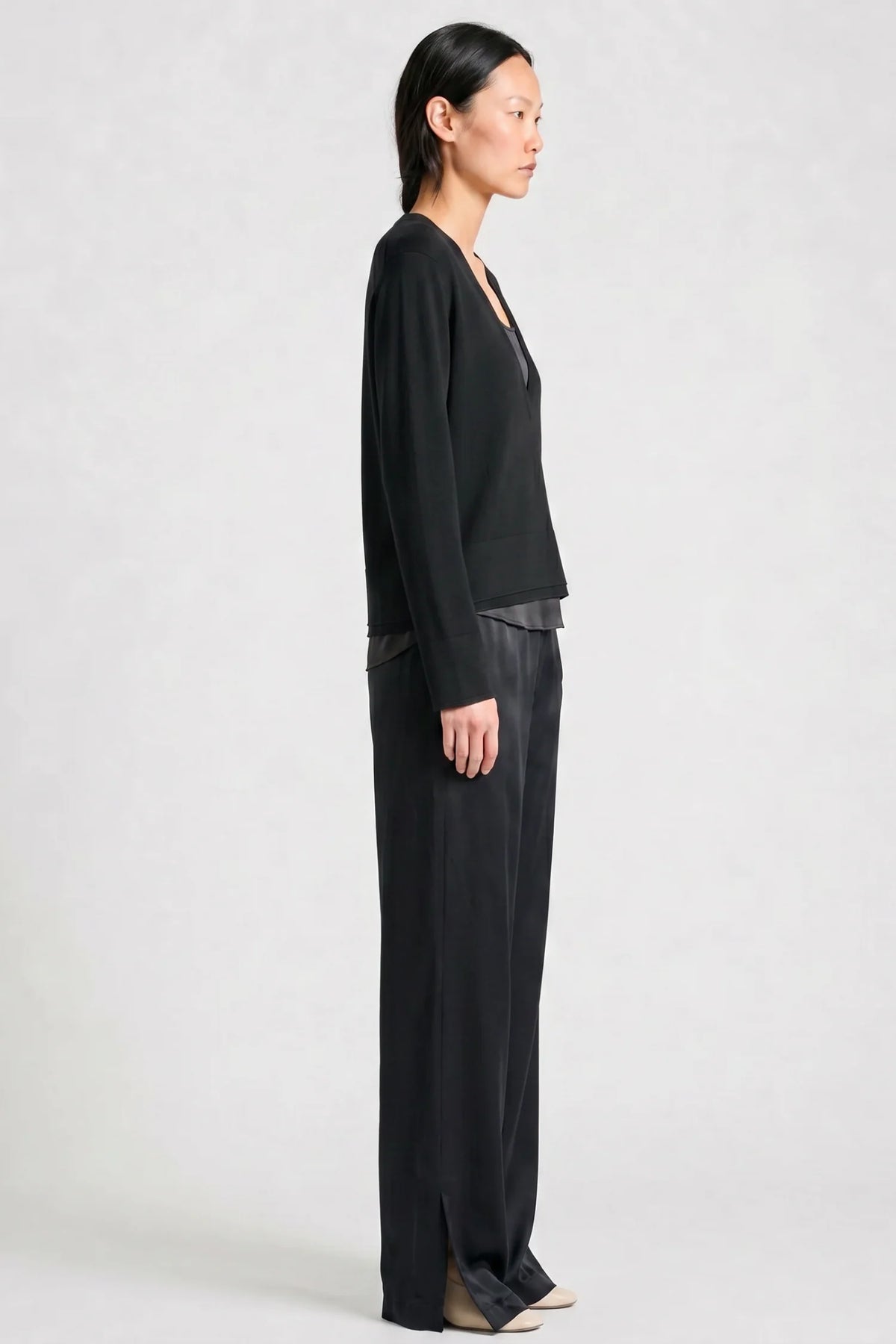 model wearing a black vneck sweater over a silky black top with black pants on a grey background, side view