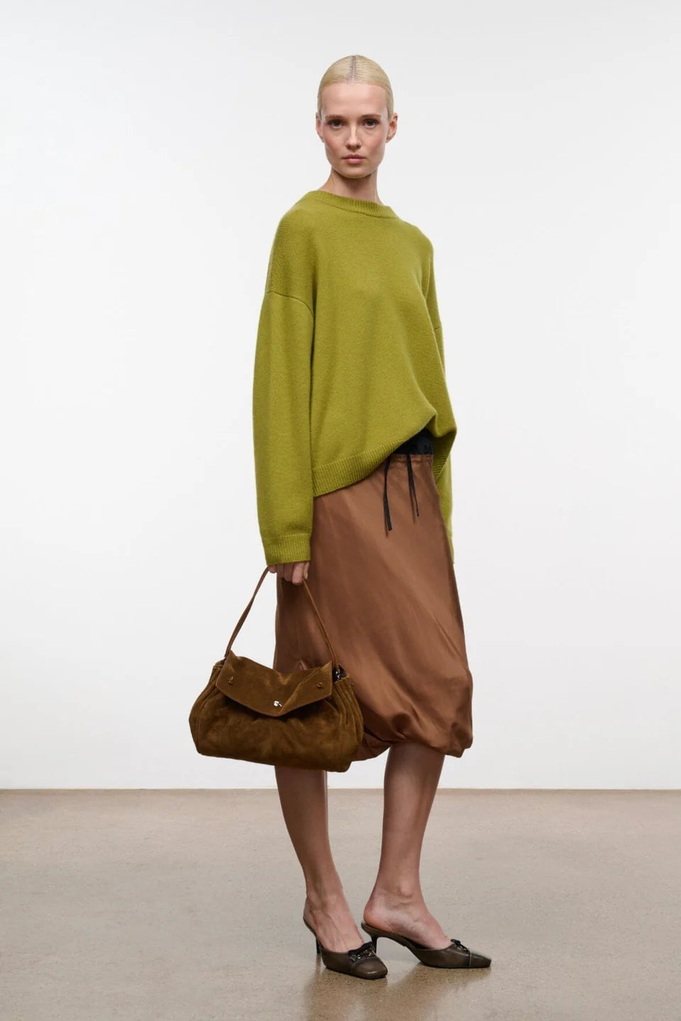 A model stands in a minimalist studio setting wearing a loose olive green knit sweater, a brown satin midi skirt, and dark brown pointed-toe mules, while carrying a brown suede flap shoulder bag.