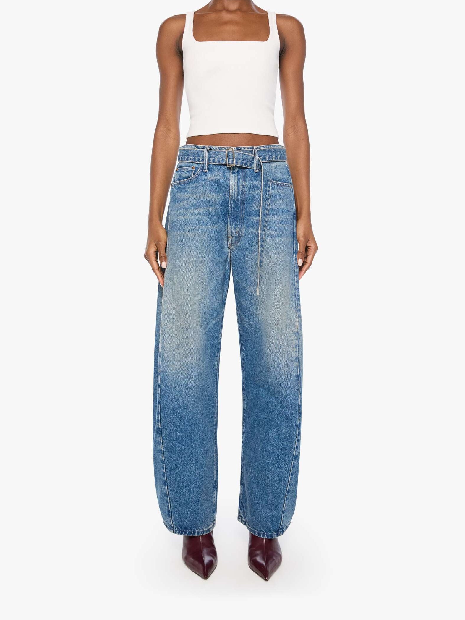 A model stands against a light gray studio background wearing a white ribbed crop top, wide-leg blue denim jeans, and dark burgundy pointed-toe heels.