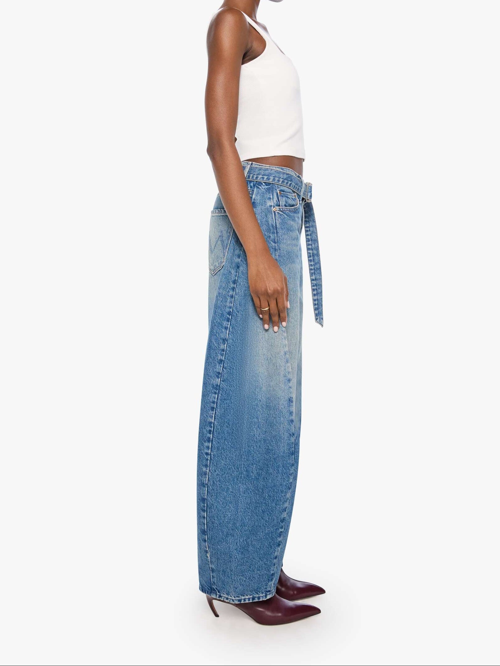 A model shown in profile wearing a white halter crop top, high-waisted wide-leg blue denim jeans with a tie waist, and burgundy pointed-toe heels against a plain light gray background.