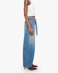 A model shown in profile wearing a white halter crop top, high-waisted wide-leg blue denim jeans with a tie waist, and burgundy pointed-toe heels against a plain light gray background.
