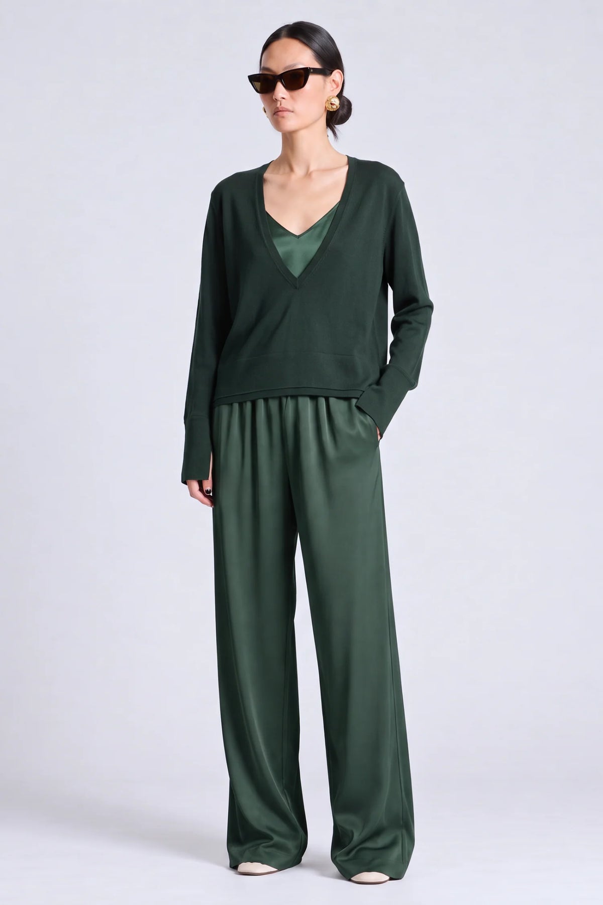 A model wearing a dark green two-piece outfit consisting of a long-sleeve wrap-style top and matching wide-leg trousers, styled with black sunglasses against a light gray studio background.