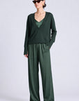 A model wearing a dark green two-piece outfit consisting of a long-sleeve wrap-style top and matching wide-leg trousers, styled with black sunglasses against a light gray studio background.