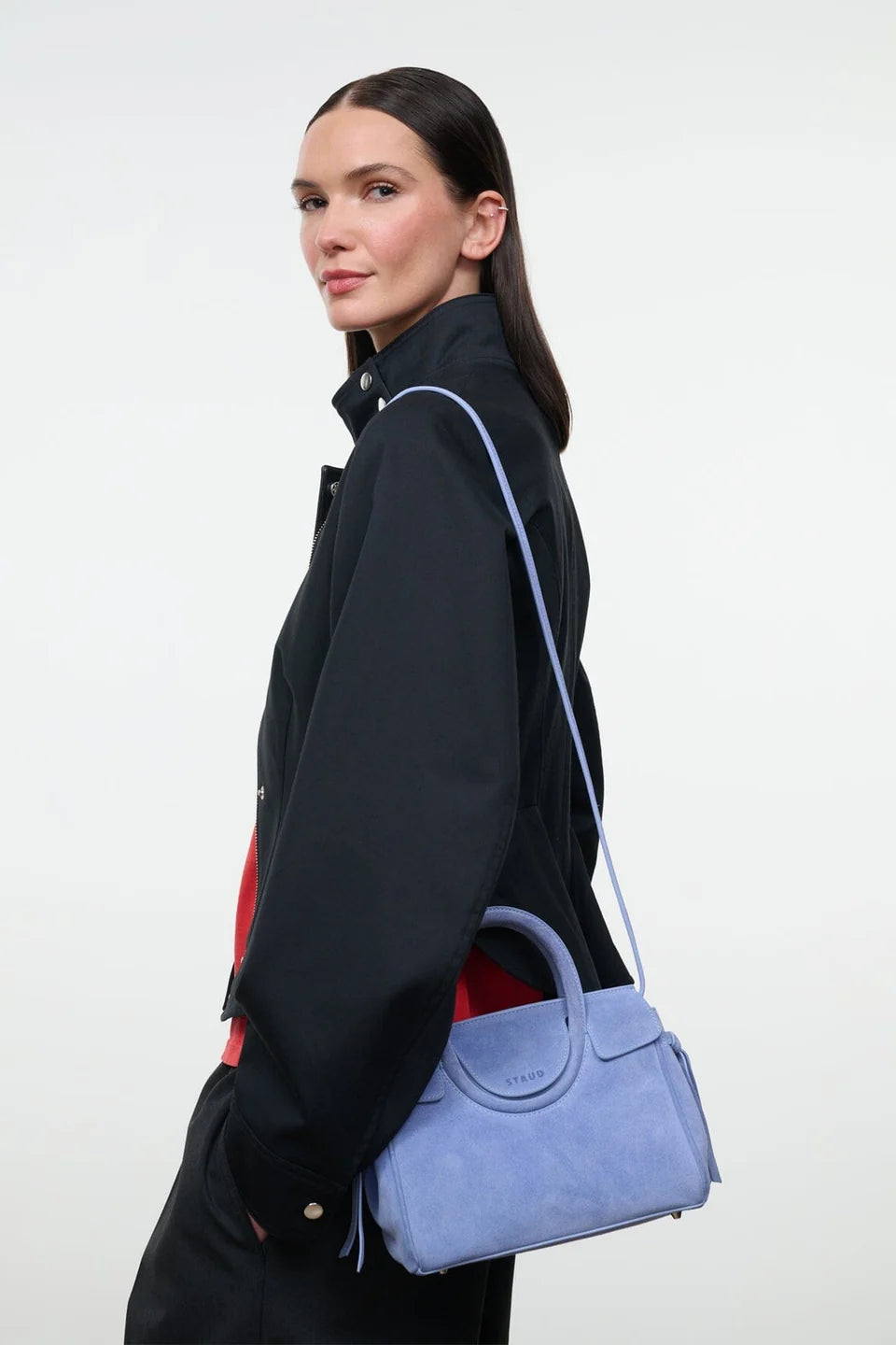 A model in a black jacket is shown from the side, carrying a light blue suede shoulder bag with a matching strap against a plain white studio background.