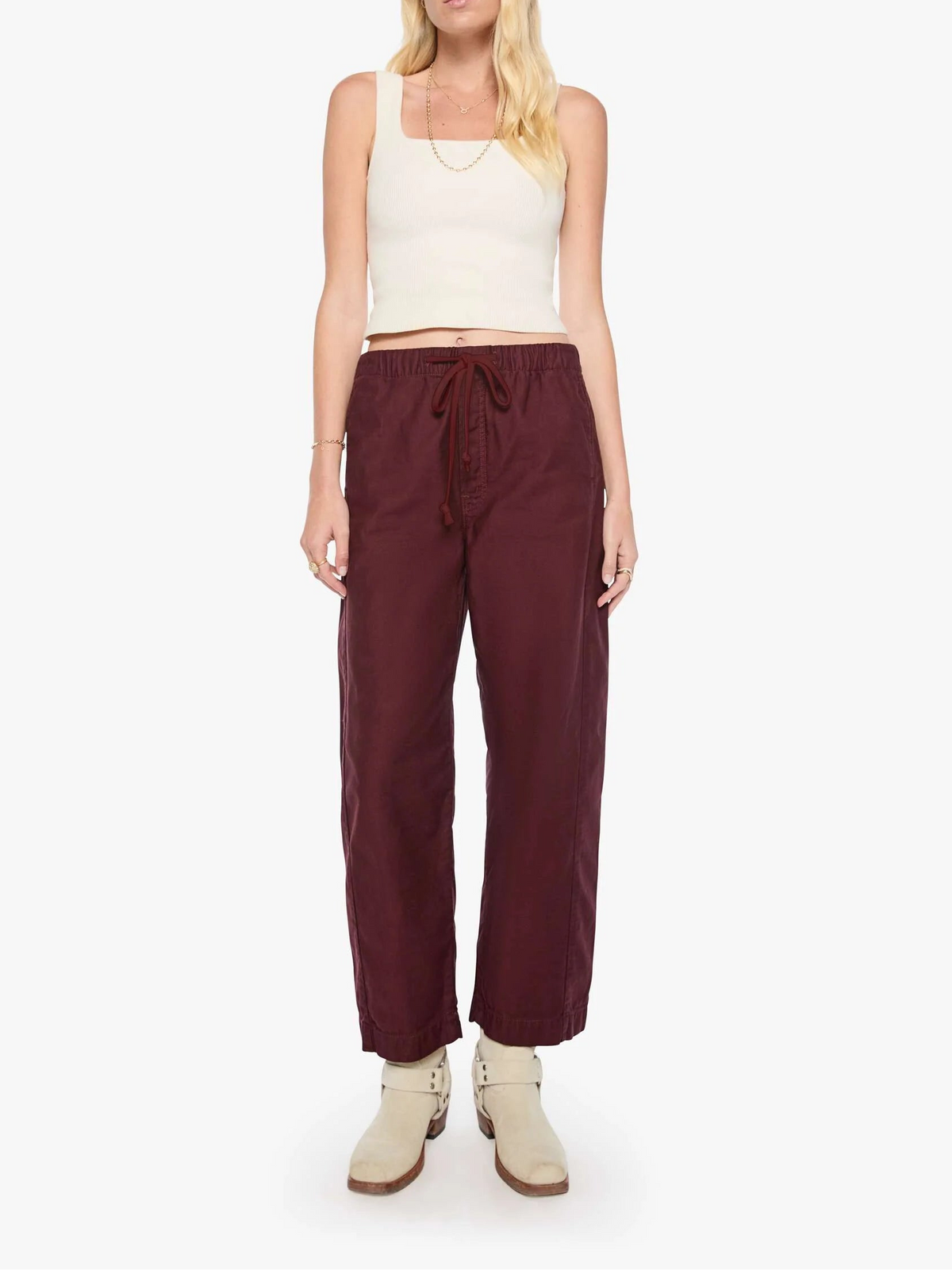 A woman modeling a pair of wide-leg burgundy drawstring pants with a cream ribbed tank top and beige ankle boots, standing against a plain white studio background.