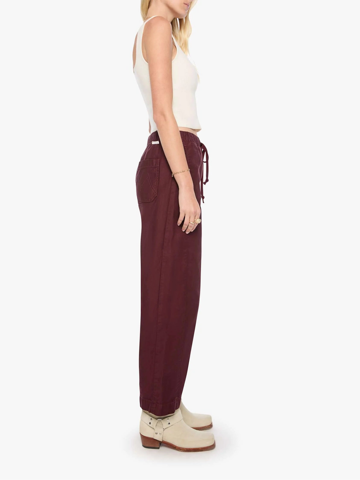 A woman shown in profile wearing loose, wide-leg burgundy drawstring pants, a white ribbed tank top, and cream ankle boots against a plain light gray studio background.