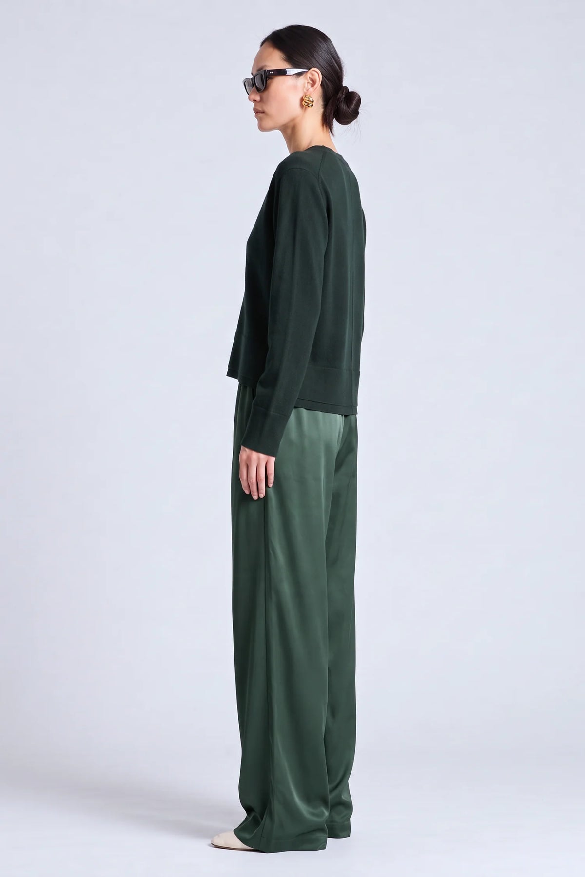 A woman in side profile wearing a dark green long-sleeve top and matching wide-leg trousers against a light gray studio background.