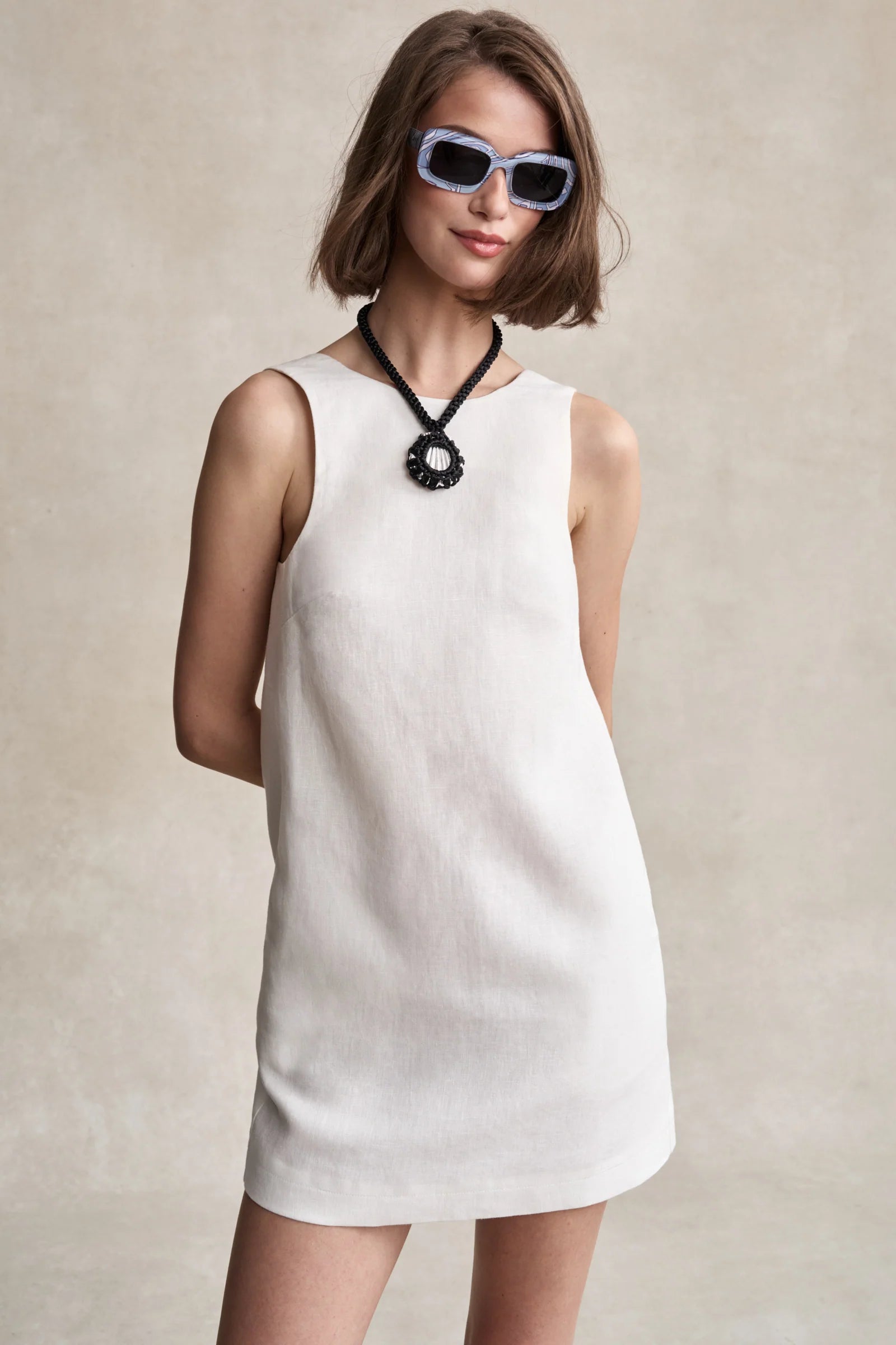 Woman wearing a white sleeveless dress with sunglasses and a necklace against a neutral background