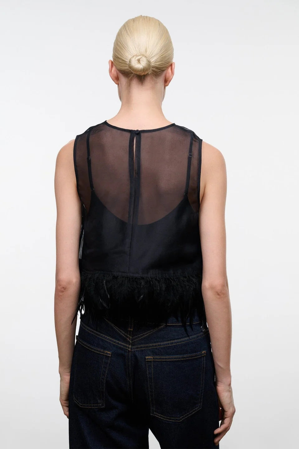A model viewed from the back wearing a black sheer mesh sleeveless top with fringe trim and dark blue jeans against a plain white studio background.