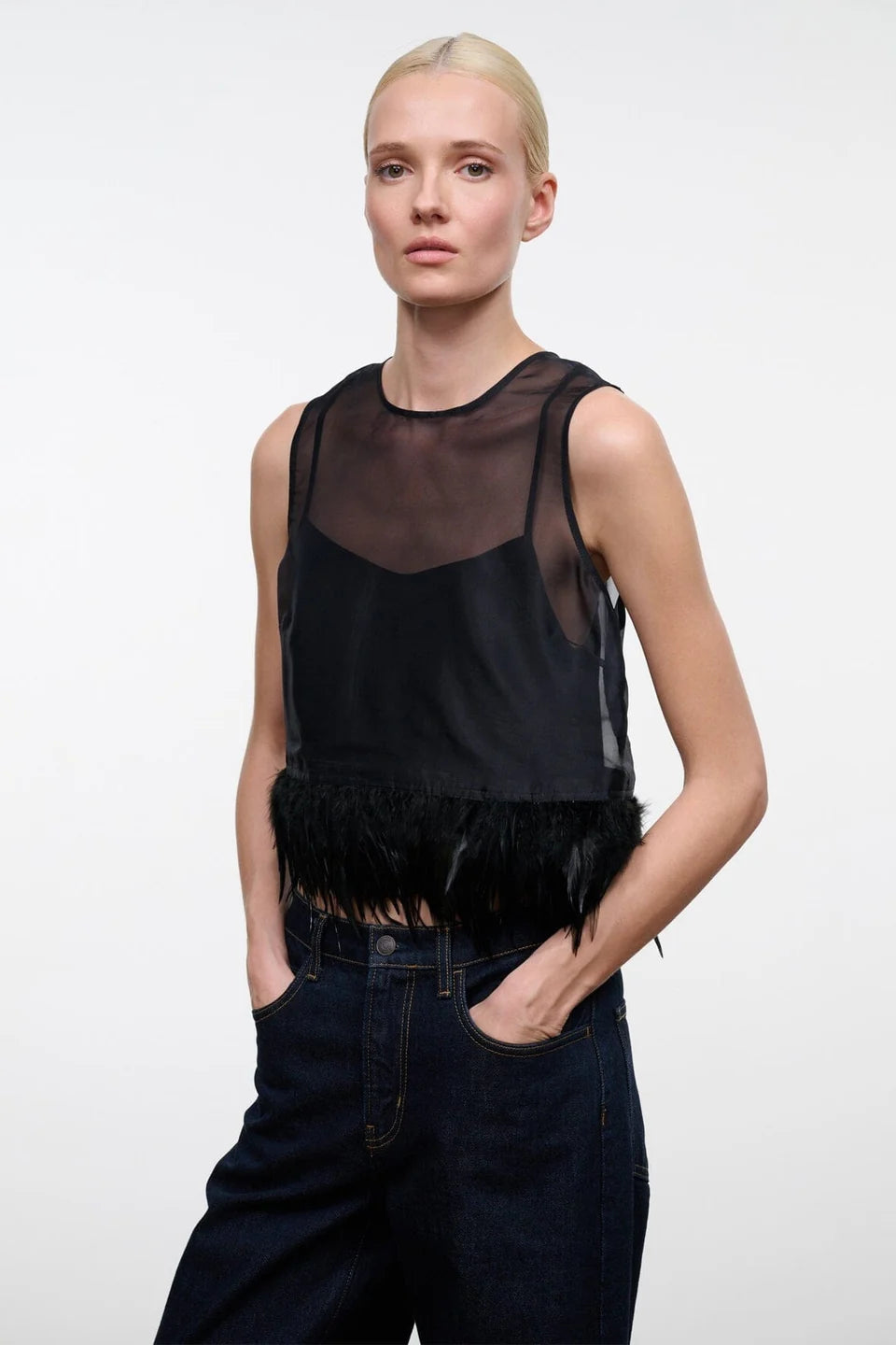 A blonde model wearing a black sheer mesh sleeveless top with a feathered hem, paired with dark blue jeans, against a plain light gray background.
